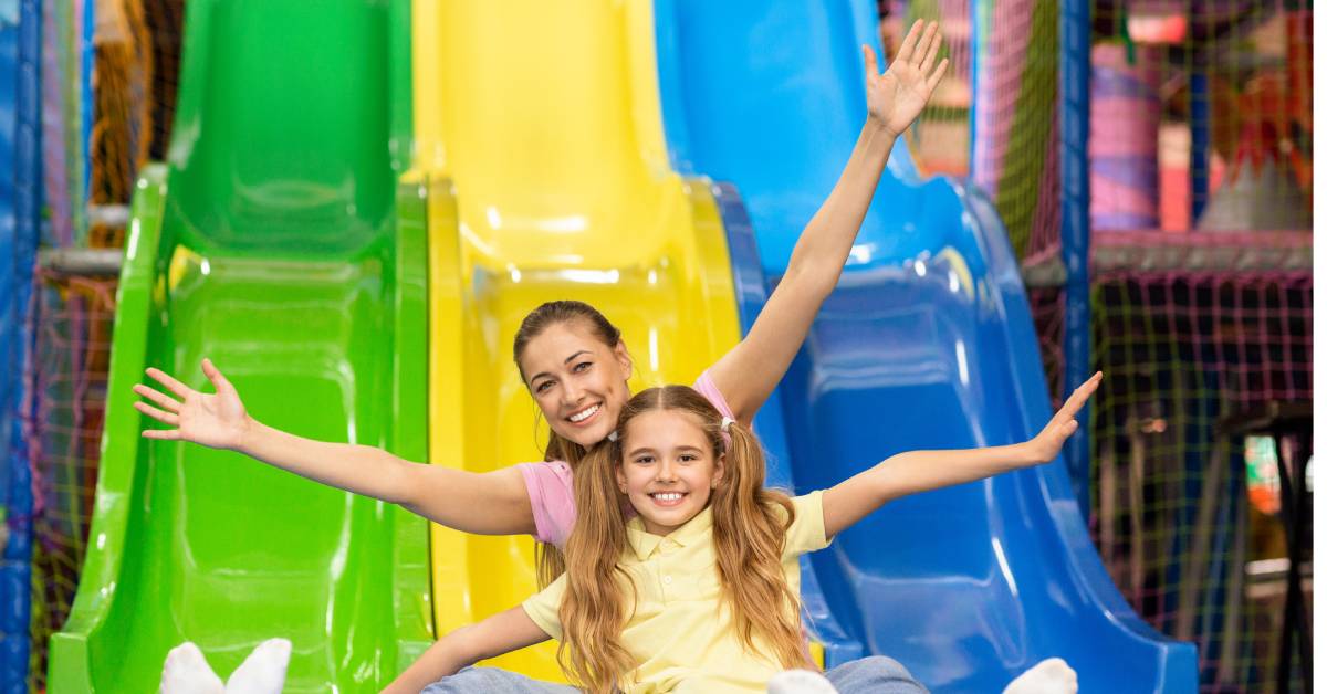 A joyful mother and her daughter smiling at the bottom of a colorful slide in an indoor children's playground.