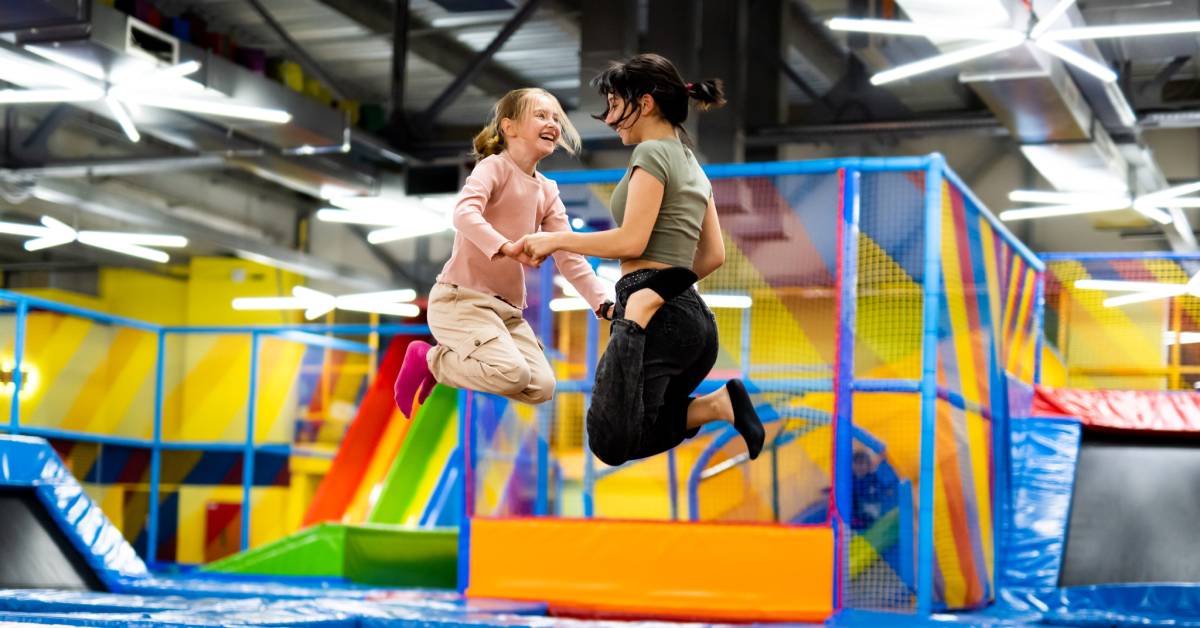 Two young girls jump on a colorful trampoline in an indoor playground. Around them are other trampolines and climbing structures.