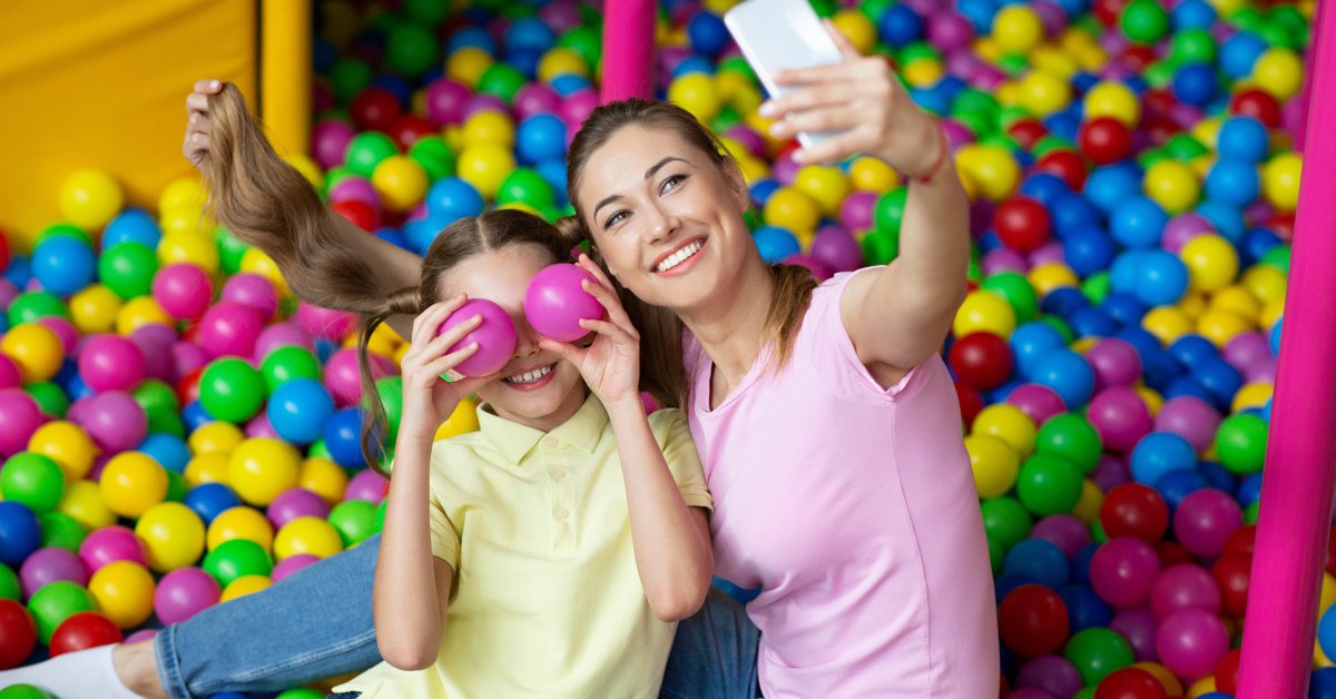 A mom and daughter in a ball pit taking a photo together with the mother's phone as the girl holds two plastic balls to her face.