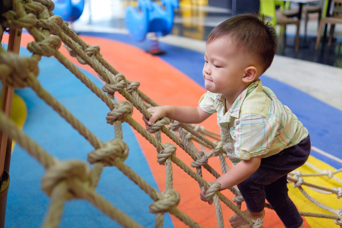 A young child in an indoor playground setting climbing the knots and rungs of a rope net with other equipment blurred behind him.