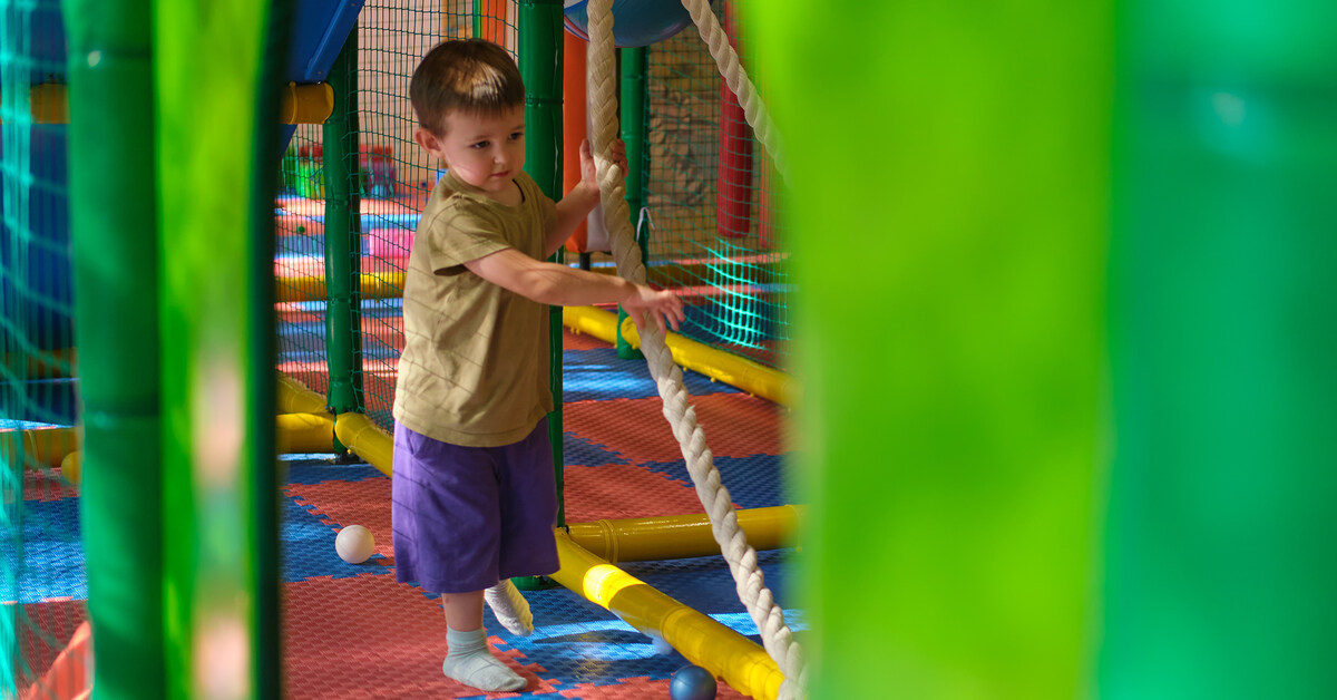 A child moving through a soft play area with foam tiles, ropes, scattered plastic balls, and walls made of soft material.