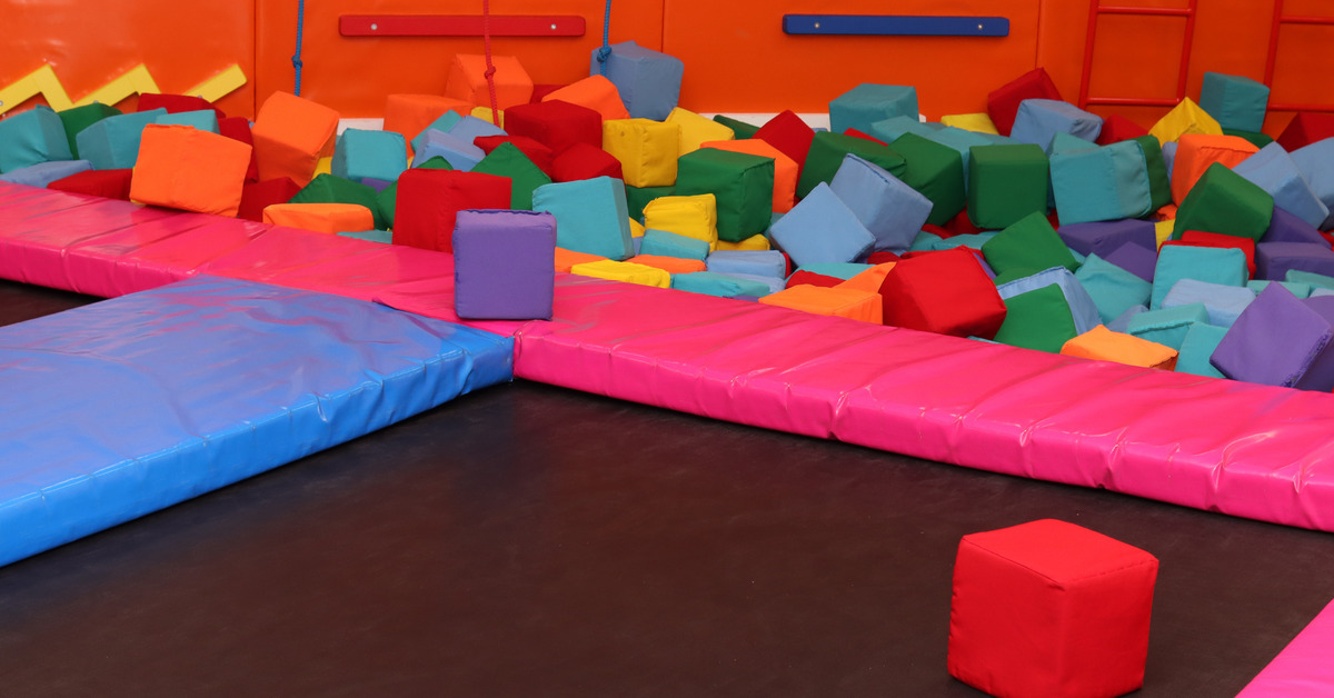 An indoor trampoline area next to a pit of soft blocks of various colors and a small climbing wall with ropes and bars.