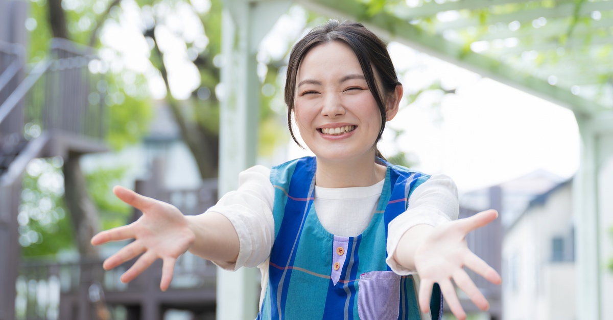 A woman in a white shirt and a blue, green, and purple smock smiles and holds out both arms in an inviting gesture.