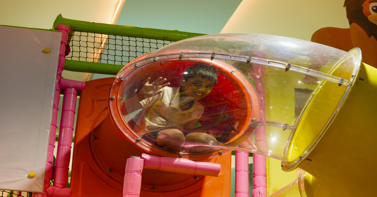 A child smiles from the inside of a clear plastic tunnel that's part of an indoor playground system.