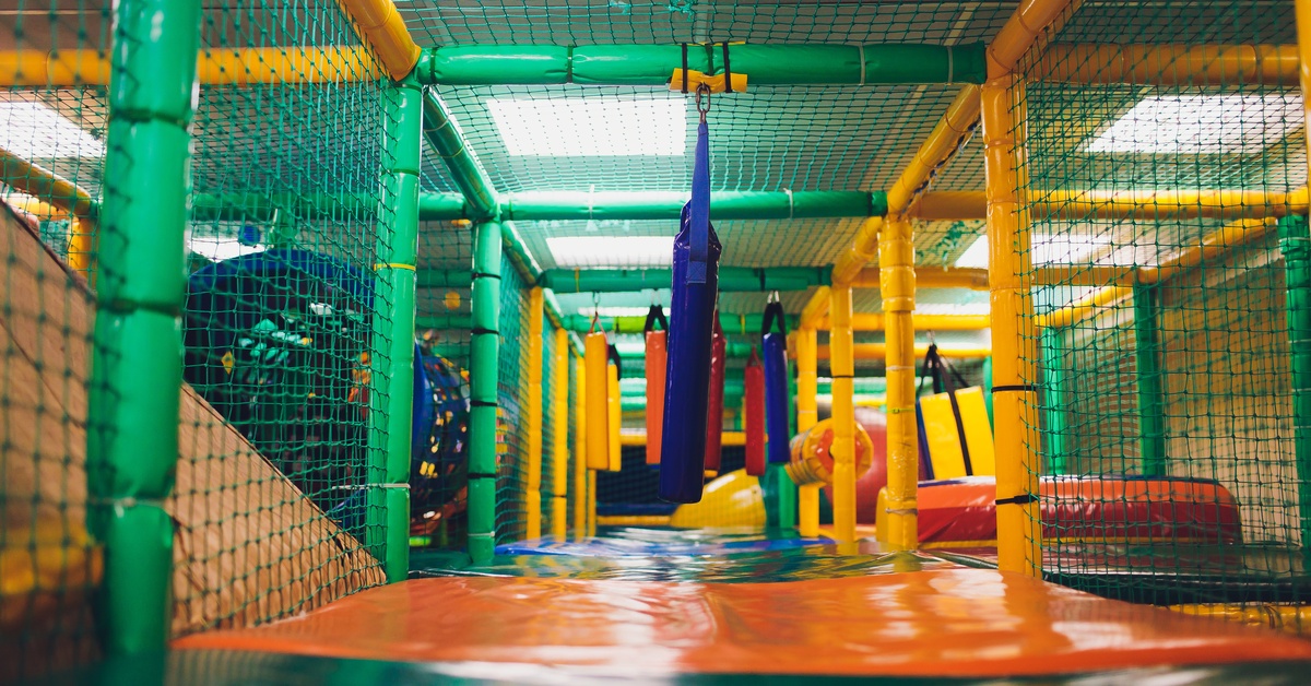 An indoor playground with bright green net walls, dangling obstacles, and soft blue and orange plastic flooring.