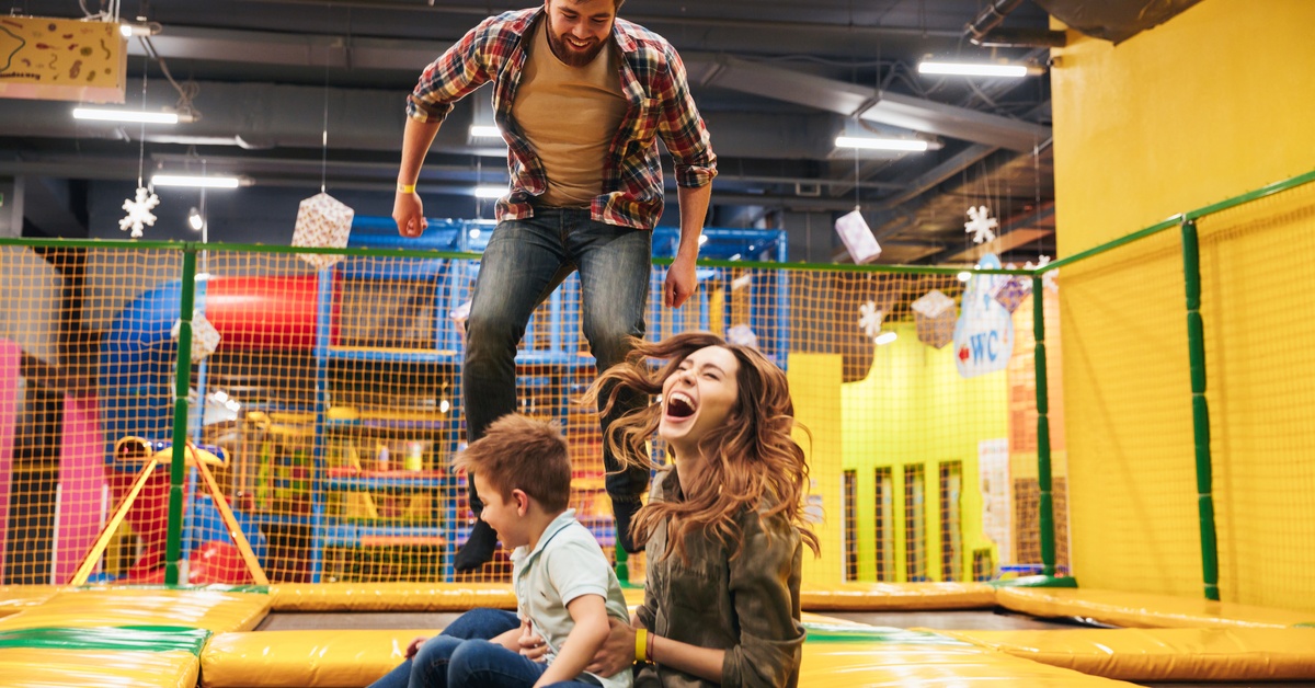 One man, one woman, and a young child bounce on the trampoline in an indoor playground. They are all smiling and laughing.