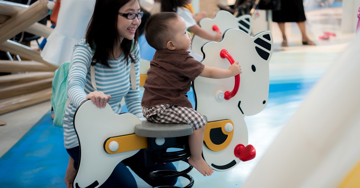 Two children, a toddler and a preteen, play together with the wooden toy horses at an indoor playground.