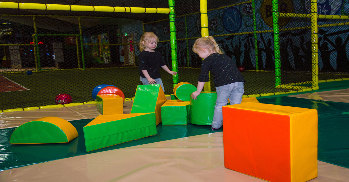 Two twin girl toddlers in similar clothing playing with large, soft building blocks in an indoor playground.