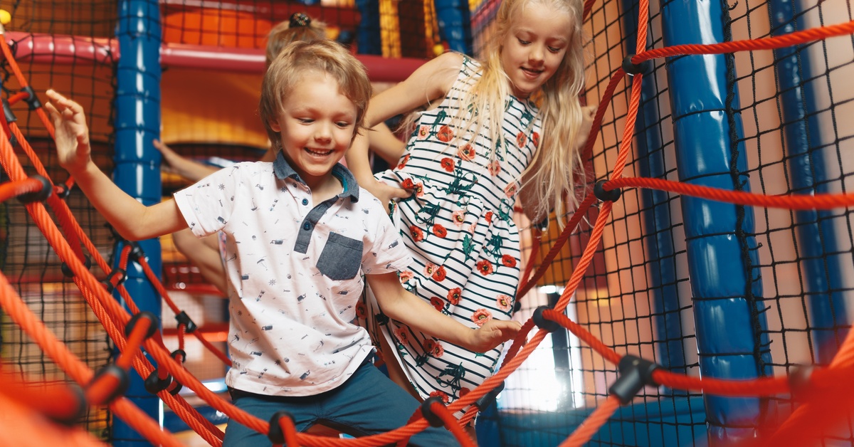 A young boy and girl climb on red ropes at an indoor playground, with black safety netting in the background.